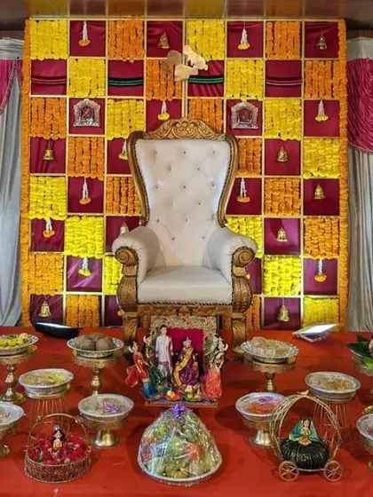 A clear, well-lit photograph of a Seemantham stage. The focus is on the checkered marigold backdrop and the neat arrangement of offering plates and dolls in front of the throne.