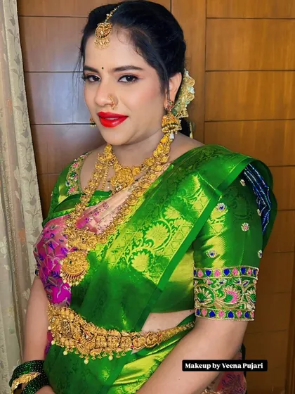 A vibrant and classic bridal look. The bright red lip and defined eyes stand out beautifully against the rich green and pink of her silk saree and heavy gold jewelry.
