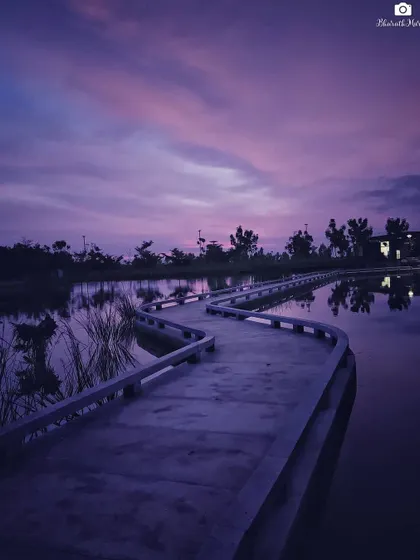 A winding concrete boardwalk reflects the beautiful purple and pink hues of the twilight sky over a calm lake. This serene landscape captures a peaceful evening in Bangalore.