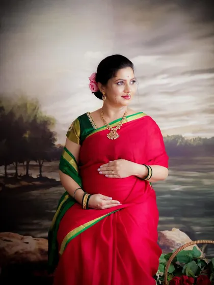 A serene and traditional portrait. The mom-to-be, in a red and green saree, cradles her bump against a classic painted backdrop.