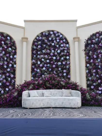 A beautiful stage backdrop featuring three arches filled with a dense arrangement of purple and blue flowers, creating a lush and elegant photo wall.