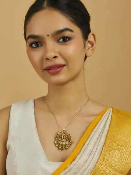 A model gracefully wears a gold-toned temple pendant with a deity motif and ghungroo bead drops. It's paired with a simple chain, showing how a single statement pendant can define a look.