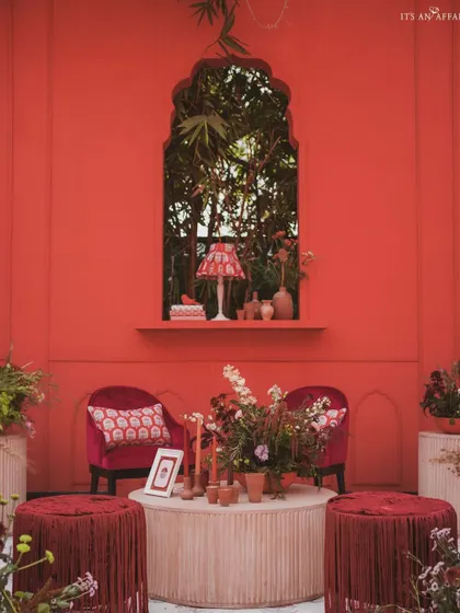 An intimate seating nook within the 'Gulal Bagh' theme, featuring a rich red backdrop, velvet armchairs, and custom-printed textiles.