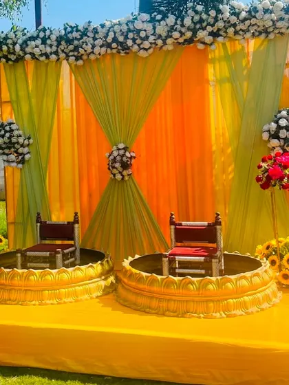 A classic outdoor Haldi setup with seating for the bride and groom. The stage is decorated with yellow and orange drapes, white floral arrangements, and sunflowers for a fresh, traditional feel.