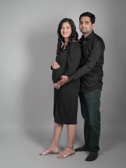 A classic couple's portrait in the studio. The expecting parents are dressed in matching black, creating a cohesive and timeless look against a simple grey background.
