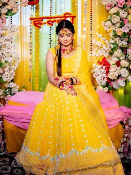 A candid moment of the bride during her Haldi ceremony, seated against the vibrant backdrop of flowers and drapes.