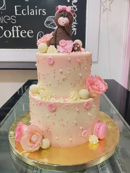 A charming two-tiered pink cake for a little one's celebration. The cake is decorated with edible pearls, pink buttons, fresh roses, and a cute fondant teddy bear topper.