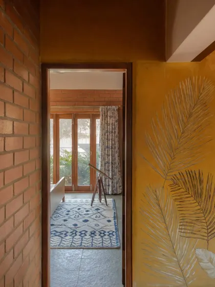 A view from the corridor into a bedroom, framing the garden beyond. The yellow oxide wall with leaf art contrasts beautifully with the exposed brick.