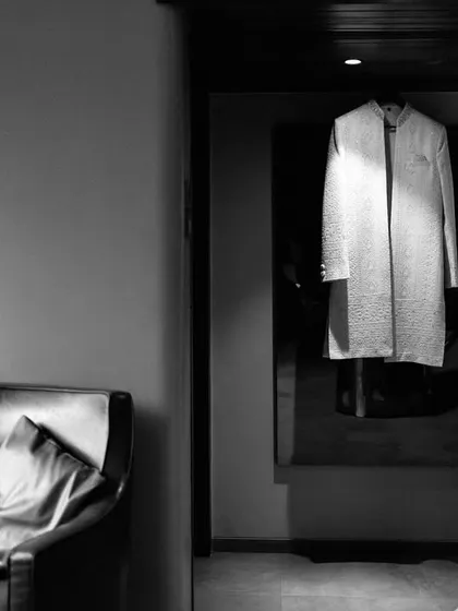 An artistic black and white shot of the groom getting ready, his sherwani hanging in the background, creating a sense of anticipation.
