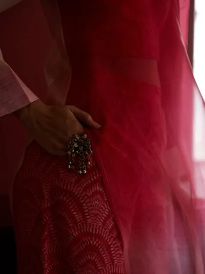 A close-up detail shot focusing on the texture of the red patterned fabric and the large, ornate ring. The sheer dupatta adds a layer of softness, creating a beautiful interplay of textures.