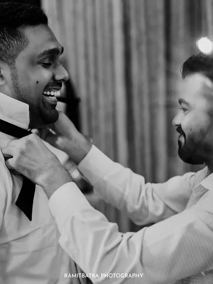 A candid black and white photo of a friend helping the groom with his bow tie, a classic getting-ready moment.