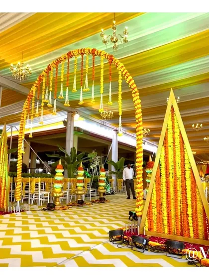 An entrance arch for a Haldi ceremony, decorated with marigold garlands and traditional floral arrangements. The yellow and white chevron flooring adds a modern touch to the traditional decor.