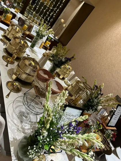 A long shot of an elegant buffet line ready for a house party. The setup includes our signature gold serving dishes, beautiful floral arrangements, and ambient lighting to create a sophisticated dining experience at home.