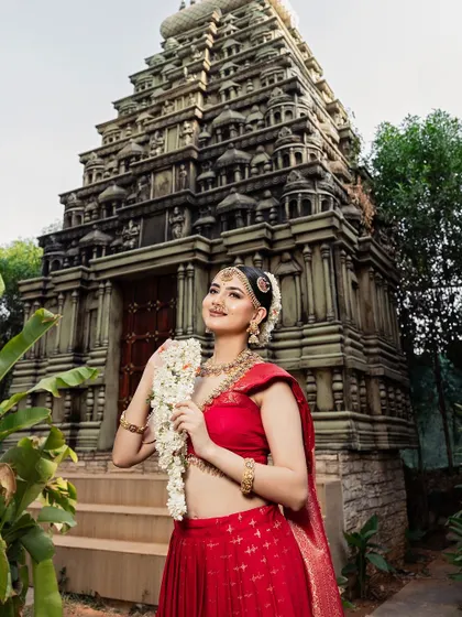 The bride stands before a temple backdrop, her red outfit and gold jewellery creating a powerful and traditional image.