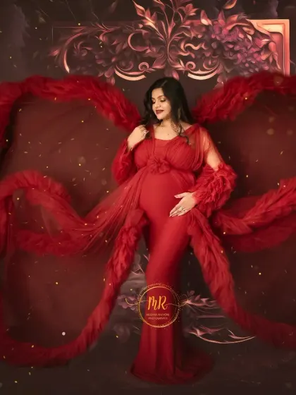 A dramatic solo portrait in a deep red gown with feathered details. The fabric is styled to create a wing-like effect against a rich, textured backdrop, perfect for a bold pregnancy announcement.