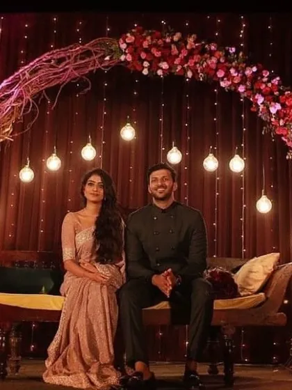 A romantic stage setup for an engagement or reception. A floral arch made of twigs and red roses frames the couple's seating area, with hanging light bulbs adding a warm glow.