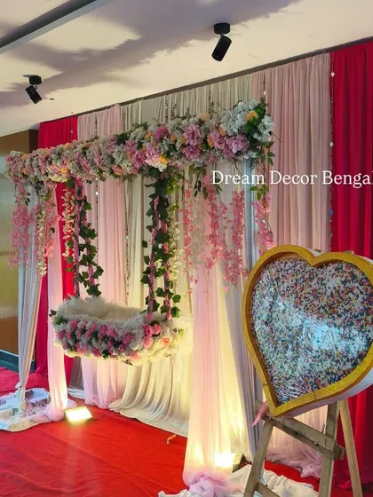 This naming ceremony decor for a baby girl uses high-quality artificial flowers in shades of pink. The hanging cradle and heart-shaped name reveal board add a sweet, personal touch.