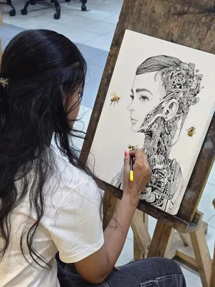 A student works on her surreal portrait, using a micron pen to draw complex mechanical details emerging from the subject's neck and head.
