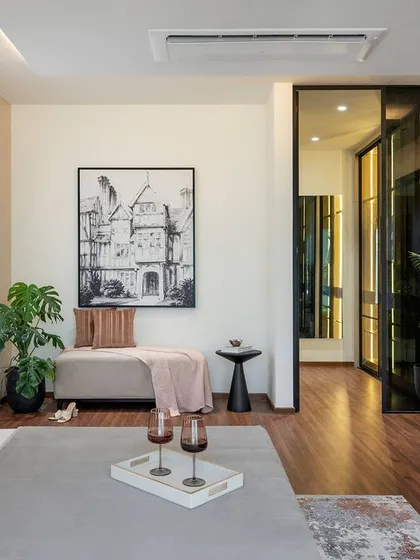 This spacious bedroom is designed as a complete personal sanctuary. It features a dedicated seating area, hardwood floors, and a glass-paneled door leading to a walk-in closet. The use of potted plants and black-and-white art adds a fresh, contemporary feel.