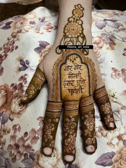 A simple and traditional Gangaur mehndi. The palm features the sacred mantra "Gor Gor Gomti" inside an ornate frame, with floral details on the fingers.