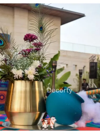 A themed centerpiece for a Krishna event, featuring a brass vase with peacock feathers, a pot of 'makhan' (butter), and a flute.