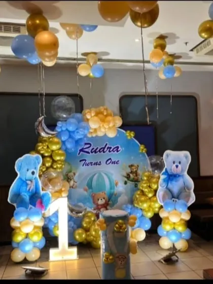A cute teddy bear themed first birthday party. The setup includes a custom backdrop with bears in a hot air balloon, large bear cutouts, and a blue and gold balloon arch.
