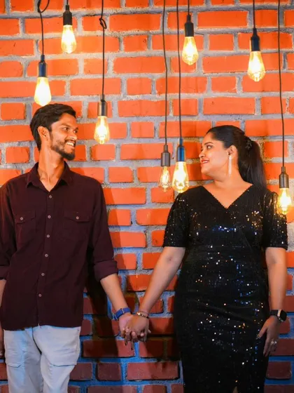 A simple and sweet pre-wedding photo of a couple holding hands against an exposed brick wall with hanging lights.