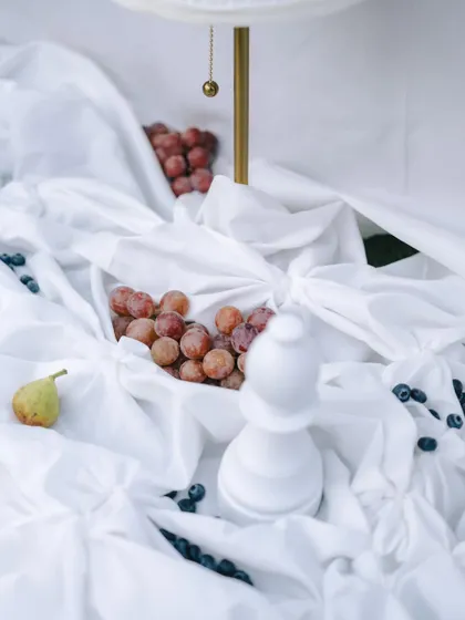 A detail shot of the layered white drapes, with a white chess piece and fresh fruits creating a still life of quiet indulgence.