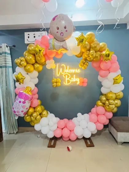 This is a modern and stylish setup for a baby shower. It features a circular balloon wreath in pink, white, and gold, with a 'Welcome Baby' neon sign that looks great in photos.