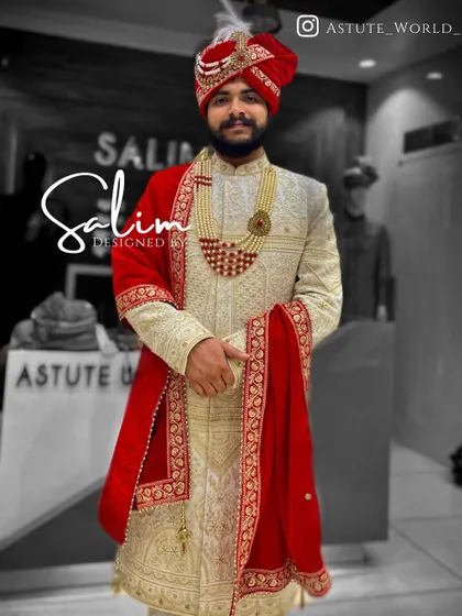 A classic groom's ensemble featuring an ivory sherwani with gold embroidery. The look is completed with a deep red safa and a matching red velvet stole, creating a timeless and regal combination.