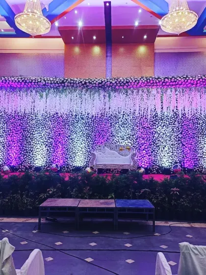 A wide shot of a large reception hall stage. The setup features a ceiling-to-floor curtain of white flowers, creating a stunning visual texture. The stage is lined with greenery, and the lighting can be customized to create different moods.