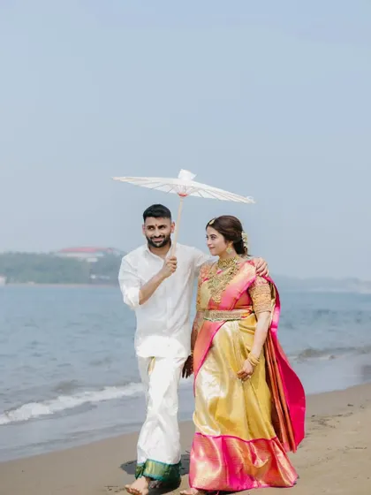 A South Indian couple walks along the beach, the groom holding a traditional white umbrella, creating a serene and picturesque moment.