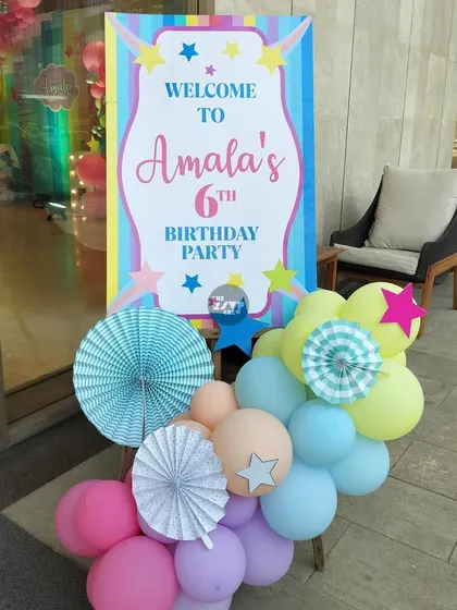 A cheerful welcome sign for Amala's 6th birthday party, decorated with pastel balloons and paper fans.