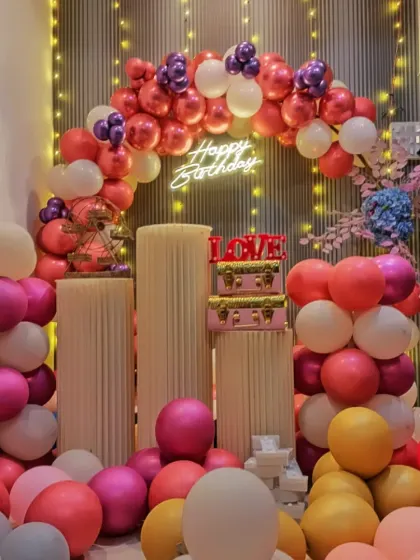 A closer look at the pink and purple birthday decoration. The setup includes plinth stands, "LOVE" blocks, and small decorative suitcases.