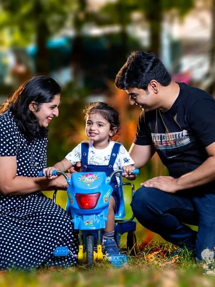 A candid family moment captured outdoors. The parents help their little one on a tricycle, sharing a laugh together in a beautiful, sunlit park.