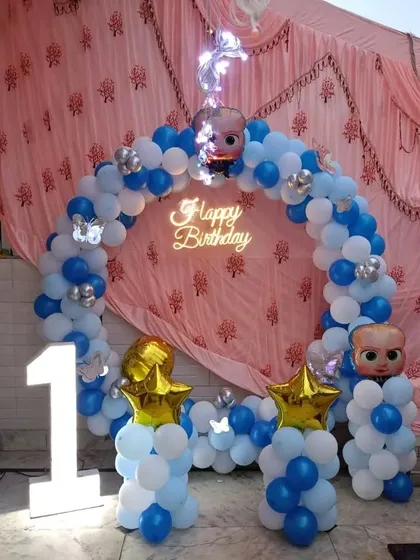 A Boss Baby themed first birthday party. This setup features a blue and white balloon arch, character foils, and a large '1' marquee light.
