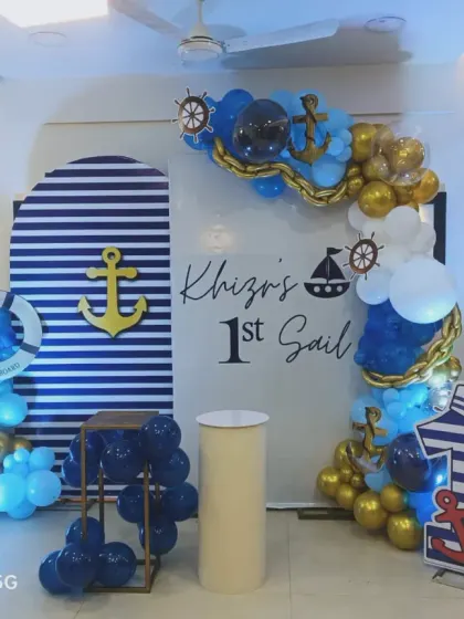 A nautical-themed 'First Sail' for a 1st birthday party. The decoration includes a striped arched backdrop, anchor props, and a blue, white, and gold balloon arch.