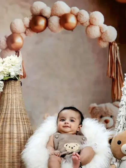 A sweet baby sits in a fluffy chair, surrounded by teddy bears and a beautiful boho-style balloon arch.