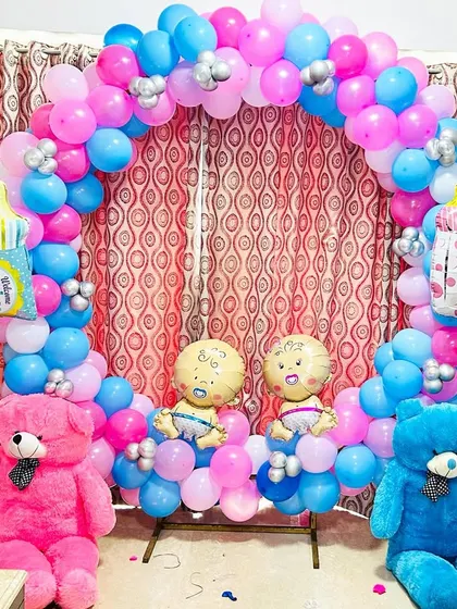 A home decoration for a baby shower, featuring a circular balloon arch in pink and blue. The setup includes teddy bears and "BABY" blocks, perfect for a gender reveal party.