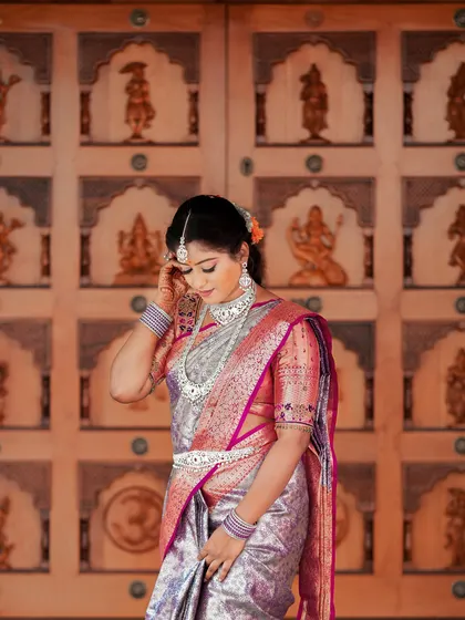 A graceful portrait of a bride in a silver Kanjeevaram saree, standing before an ornate, traditional wooden door.