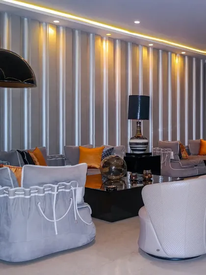 This living space features our custom-designed sofas with a unique laced detail on the arms. The soft grey upholstery is energized by vibrant orange accent pillows, all set against a backdrop of a fluted, metallic-finish wall panel.