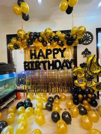 A classic black and gold home birthday decoration. This setup includes a balloon arch over a gold fringe curtain, with balloons scattered on the floor.