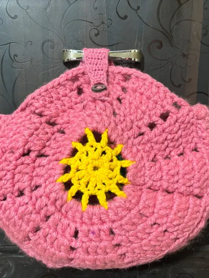 A round, pink crochet potli or pouch with a sunny yellow design in the center, featuring a metal clasp for secure closure.