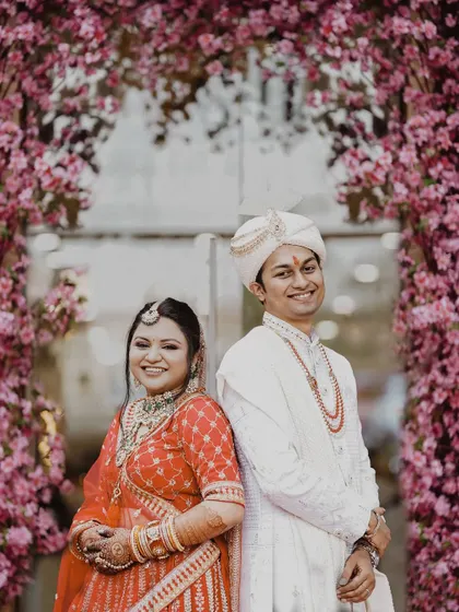 The couple stands back-to-back, framed by a beautiful pink floral archway. Their happy smiles and traditional attire make for a classic and joyful wedding portrait.