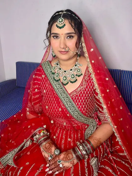 A full view of the bride seated, looking serene and beautiful. The makeup, featuring emerald green jewelry, adds a touch of regal elegance to her look.
