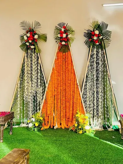 A modern and minimalist Haldi backdrop with three A-frame structures decorated with strings of marigold and jasmine flowers. The artificial grass flooring adds a touch of green.