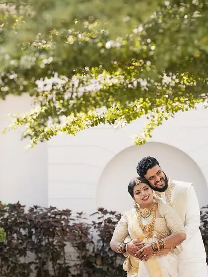 A happy, sun-kissed portrait of the couple in their white and gold wedding outfits, framed by the leaves of a tree.