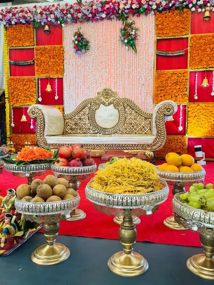 A festive Seemantham stage with a backdrop of orange marigold panels and a shimmering central curtain. The foreground is filled with beautifully arranged offerings in traditional brassware.