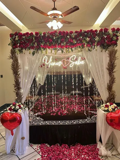A romantic wedding night setup with a bed covered in rose petals and framed by a floral canopy. Red heart-shaped balloons add a sweet touch.