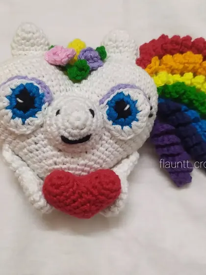A close-up of the unicorn heart plushie. It has bright blue eyes, a rainbow mane, and holds a little red heart. It's full of color and love.